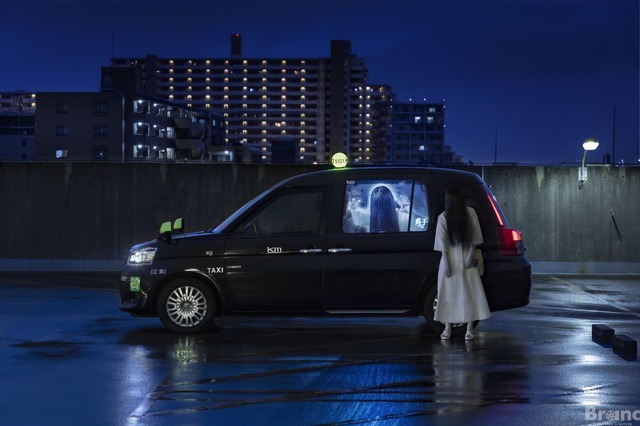 Một hình ảnh quảng cáo sắp tới "Sadako Taxi" sự hợp tác có Sadako ma quái tạo dáng bên chiếc xe taxi chủ đề Sadako ở Tokyo vào ban đêm.